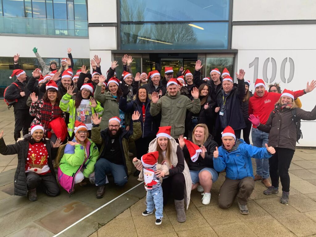 A group of more than 30 people, some in xmas hats, standing in a group and waving at the camera
